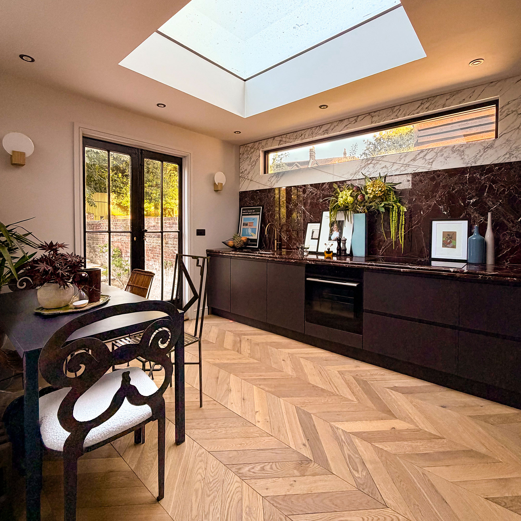 Kitchen dining space with skylight, wooden chevron flooring, mixture of chair shapes, doors to garden, dark red cabinetry, red countertops and splashbacks topped with white marble
