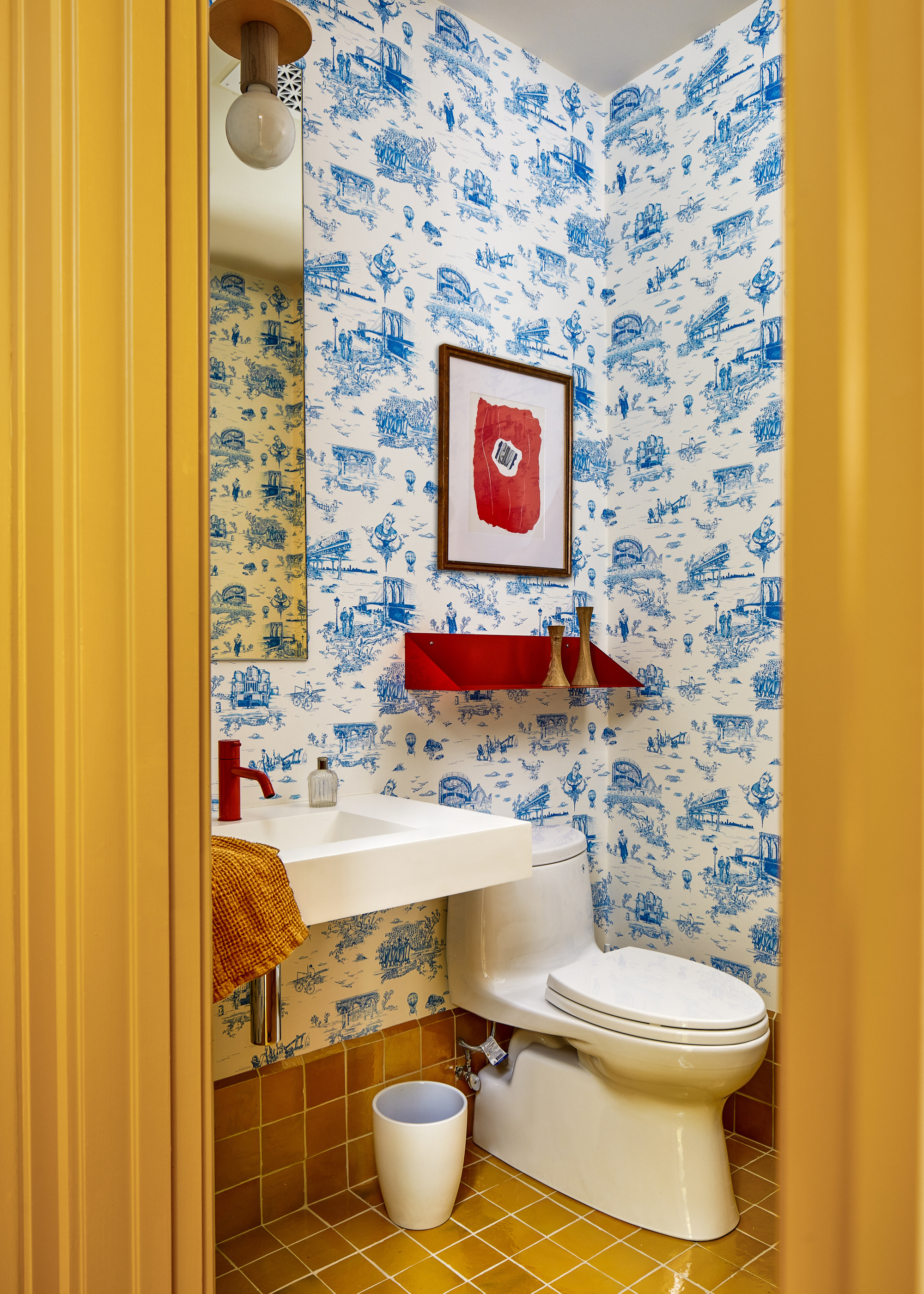 Image of a powder room with mustard yellow tiles and trim, and a blue and white patterned wallpaper. There is a red floating shelf above the toilet and the sink has a red faucet