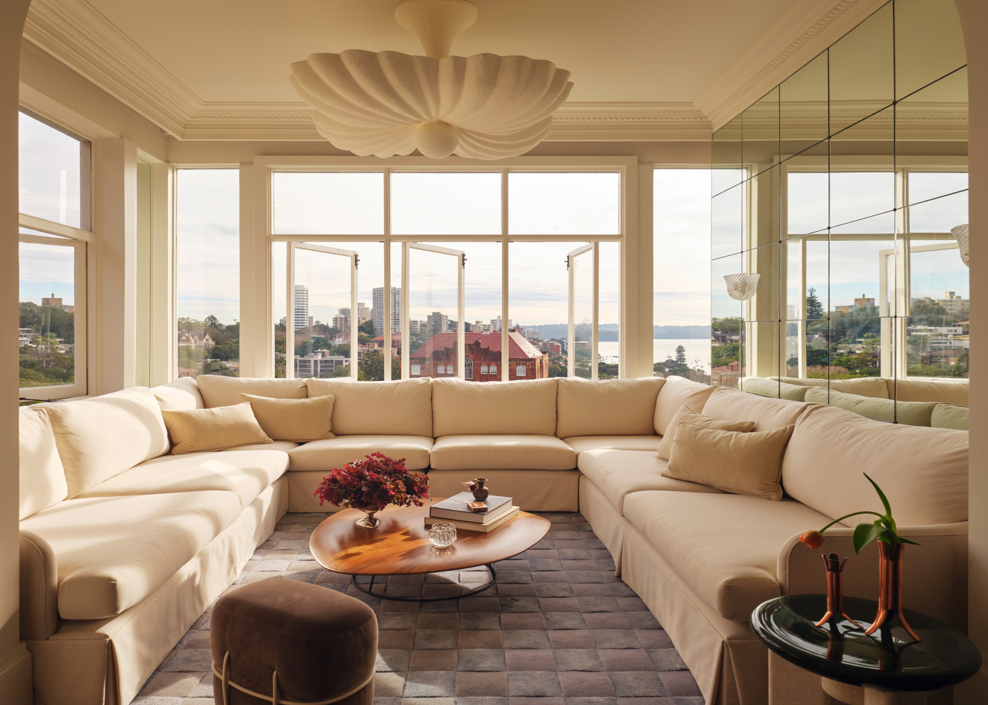 A white living room with a u-shaped sectional couch, a low coffee table, and a brown stool surrounded by windows