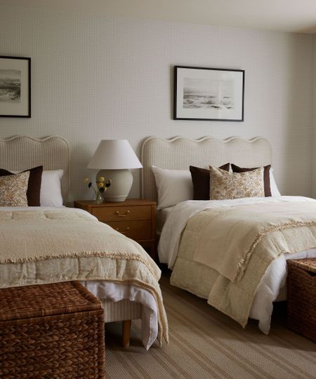 Two beds with scallopped headboard, matching white and cream bedding and brown and floral thorw pillows. Faint checkered wallpaper with framed pictures hung above beds. Bedside table inbetween