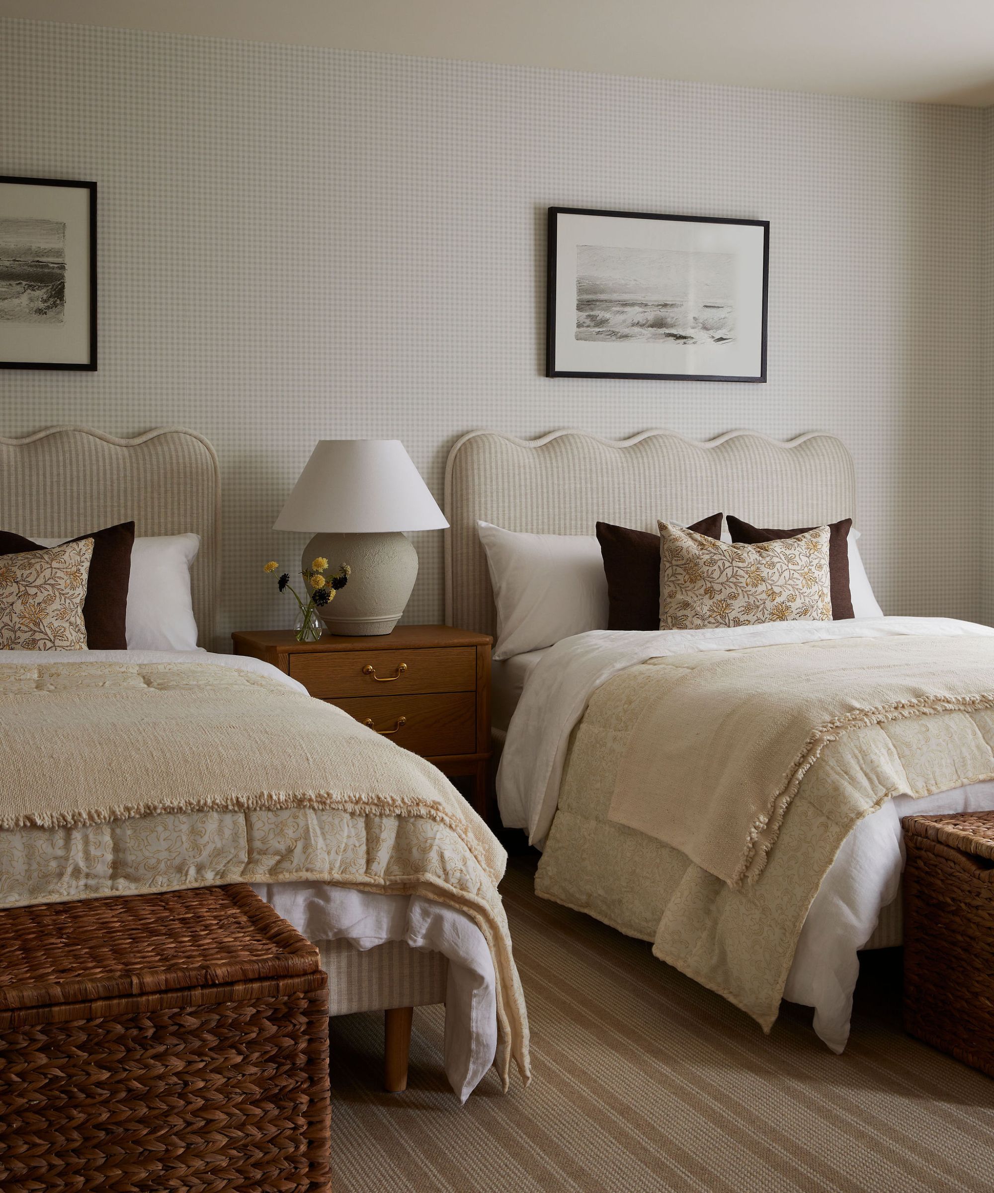 Two beds with scallopped headboard, matching white and cream bedding and brown and floral thorw pillows. Faint checkered wallpaper with framed pictures hung above beds. Bedside table inbetween