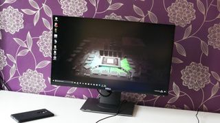 A photo of the Dell S2417DG pc monitor on a white desk next to a HP Elite X3 smartphone in front of a purple and gold patterned wall.