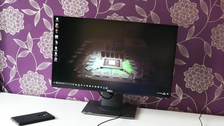 A photo of the Dell S2417DG pc monitor on a white desk next to a HP Elite X3 smartphone in front of a purple and gold patterned wall. 
