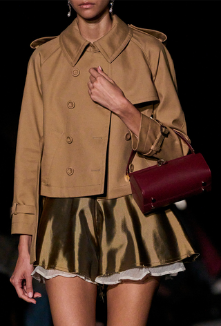 The frame bag trend is shown in a close photo from the Altuzarra spring 2024 runway show with a model wearing a cropped trench coat over a satin mini dress with a red frame bag