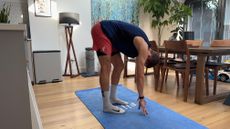 Sam RIder performs an Elephant Walk stretch on a mat in his dining room. He is standing, bent forward, hands stretching for the floor, with one leg straight and one leg slightly bent.