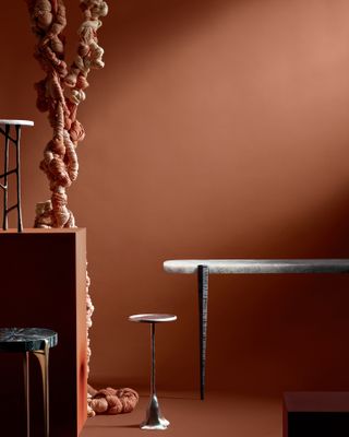 View of the Forma Side Table, Tana Side Table and Bind Console in a room with a dark terracotta floor, plinth and walls. There is also a tall dual coloured knotted structure made from fabric