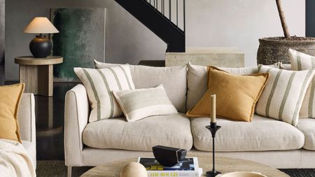 A cream sofa with cream and yellow throw cushions in a neutral living room. The edge of a round, light wooden coffee table in front, and a round side table with a black lamp on in the background. 