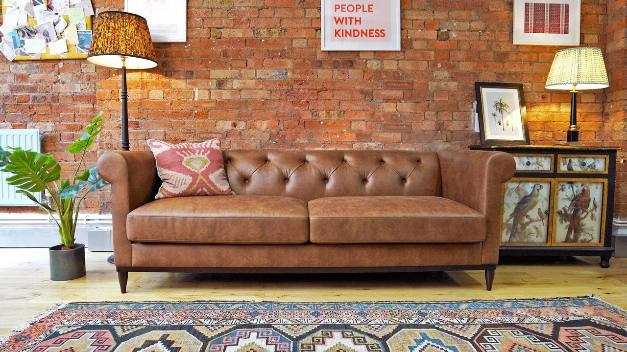 Swyft Chesterfield sofa in leather, in a living room with a lamp, rug, and a brown brick wall behind