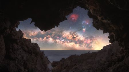 View of a starry Milky Way galaxy with colorful nebulae seen through a rocky cave opening overlooking the sea at twilight