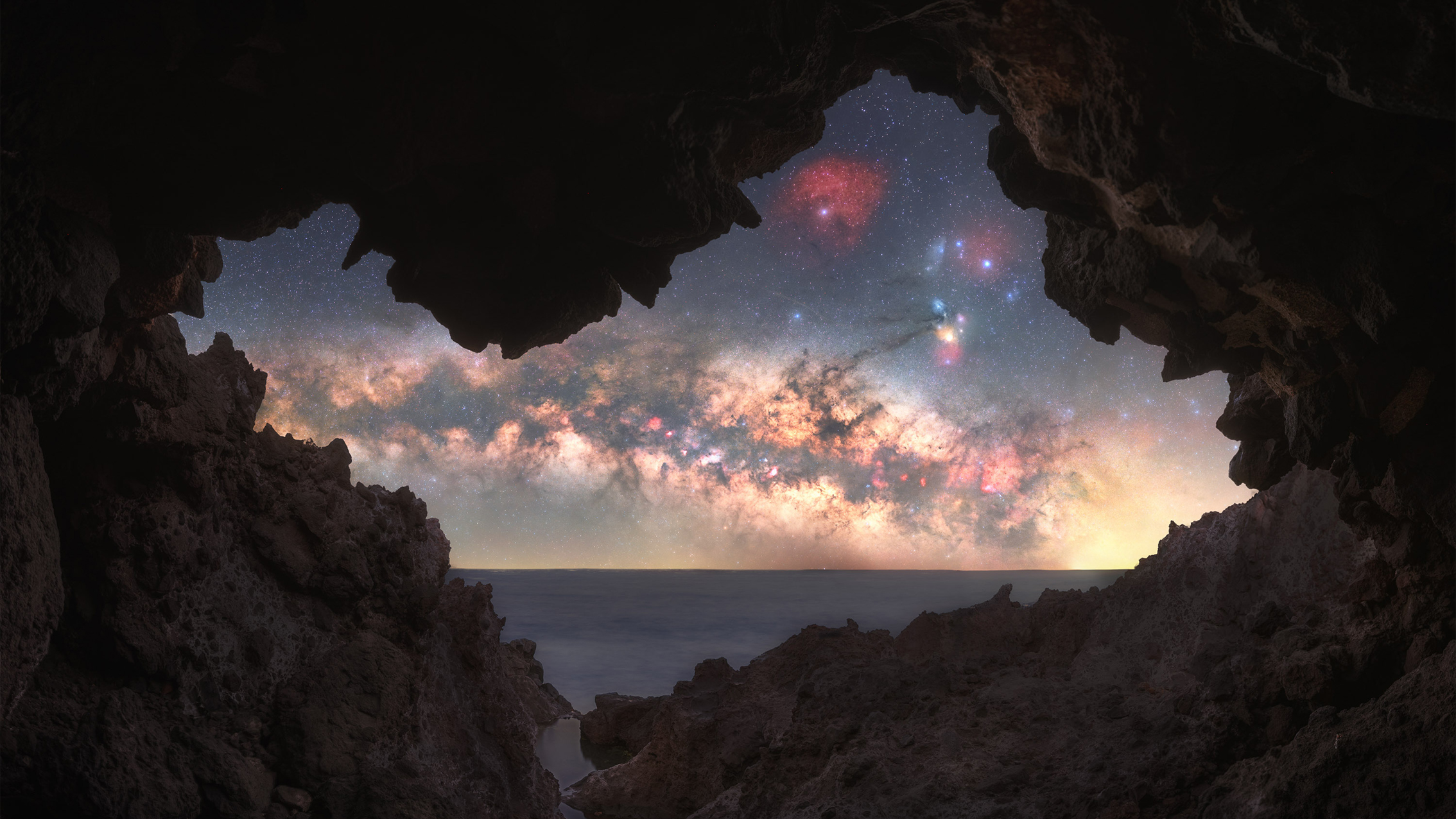 View of a starry Milky Way galaxy with colorful nebulae seen through a rocky cave opening overlooking the sea at twilight