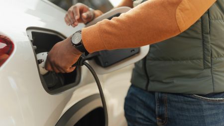 A man wearing an orange long sleeve, green puffy vest, jeans and a black and white watch plugs a charging cord into the side of a white electric vehicle
