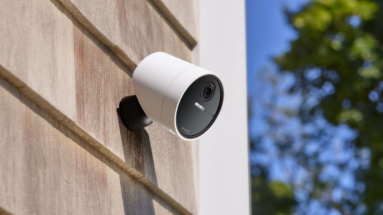 SimpliSafe wireless outdoor security camera on the side of a house in daytime with blue skies and green shrubs in the background