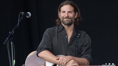 Bradley Cooper poses with a guitar on his lap and microphone on a stand.