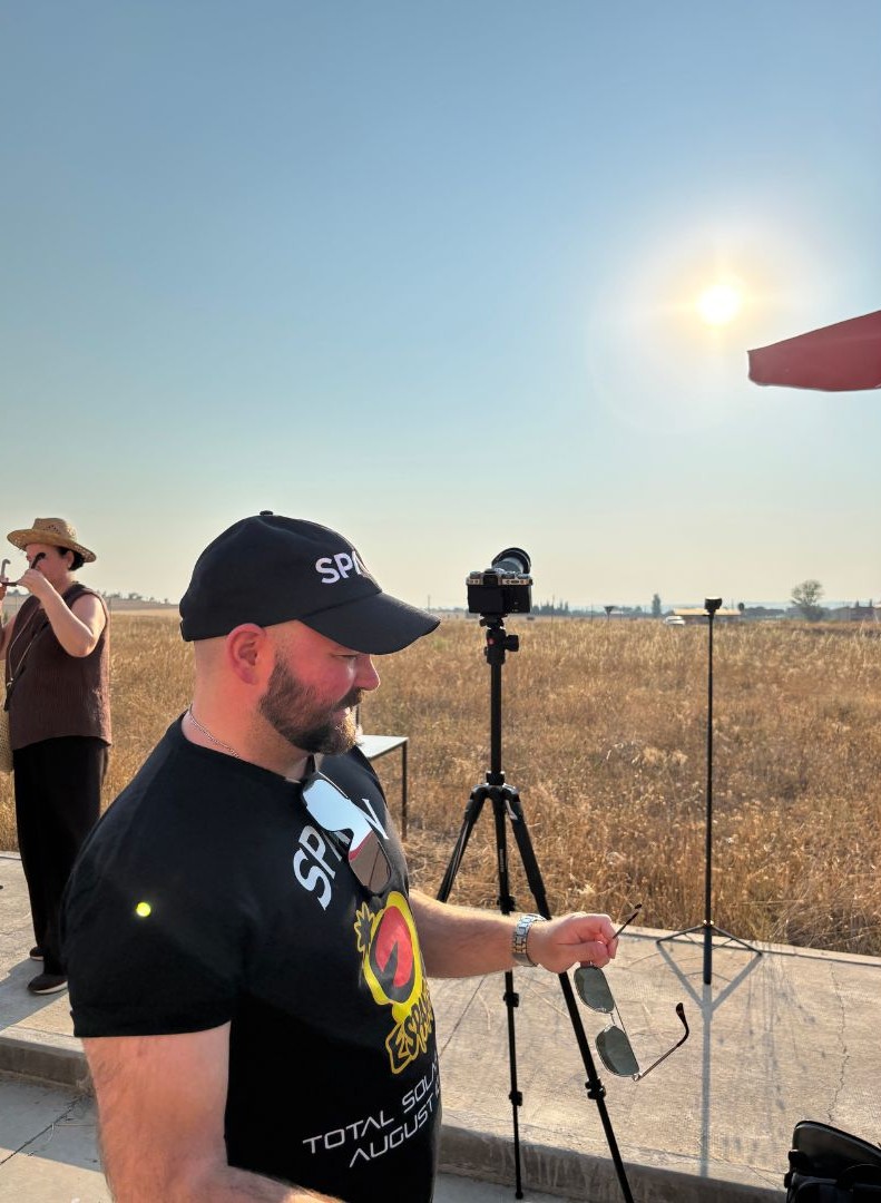 A man is standing in front of a camera with the sun shining in a blue sky above a flat field.