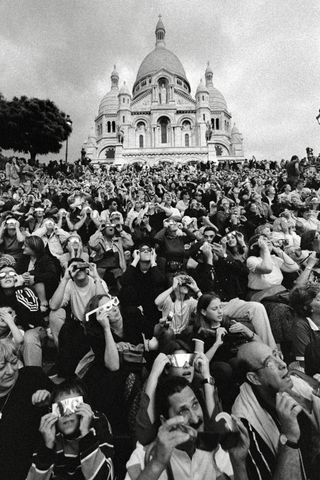 A black and white photo of lots of people looking up at a solar eclipse in front of a dome-like structure.