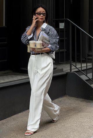 a photo showing what shoes to wear with wide-leg pants with a woman walking on the sidewalk wearing a blue and white striped button-down shirt tucked into white wide leg pants styled with white strappy heeled sandals