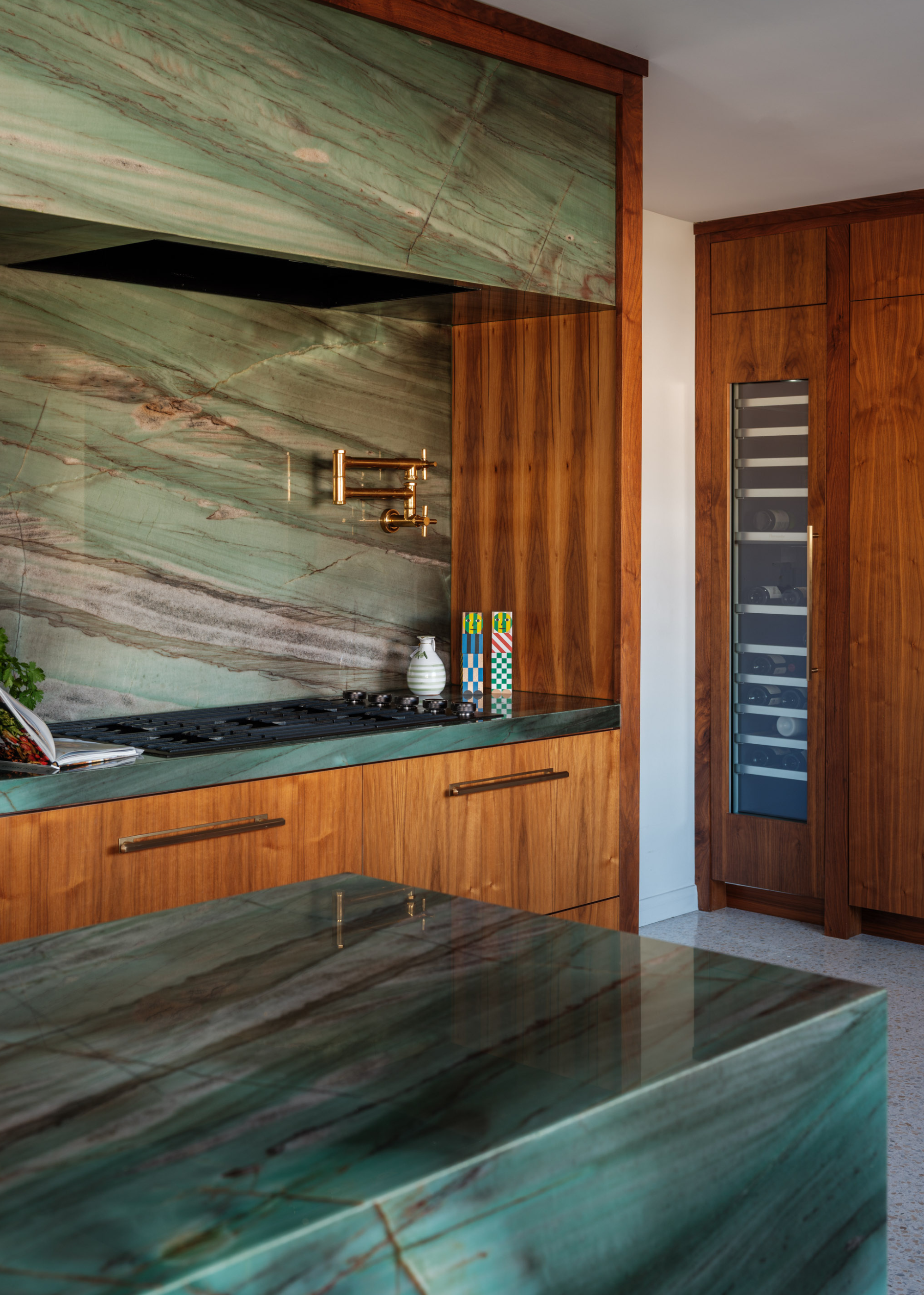 A kitchen with a blue-green marble island and matching counter and backsplash with an extendable tap by the stove, alongside a pair of colorful salt and pepper grinders, a vase and an open cookbook