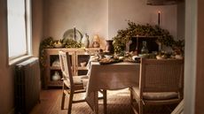 A rustic dining room with a fall garland over the mantelpiece. A rectangular table draped with a cream linen tablecloth, and wooden chairs pulled up to it. A display cabinet in the background with dinnerware inside. 