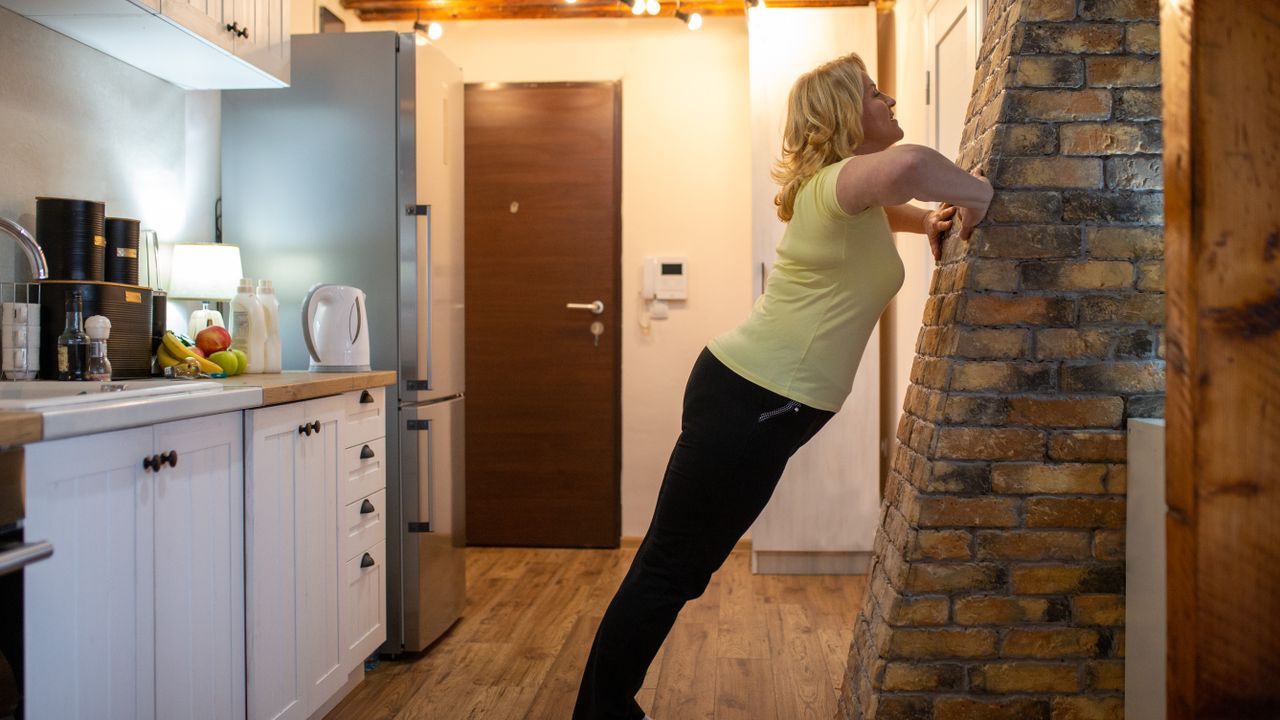 A woman uses a slanted wall in her kitchen to exercise. She is leaning towards the wall, hands resting on it with elbows bent; the rest of her body is held in a straight line from her shoulder to her feet. Behind her we see a kitchen counter with utensils, pots and fruit. 