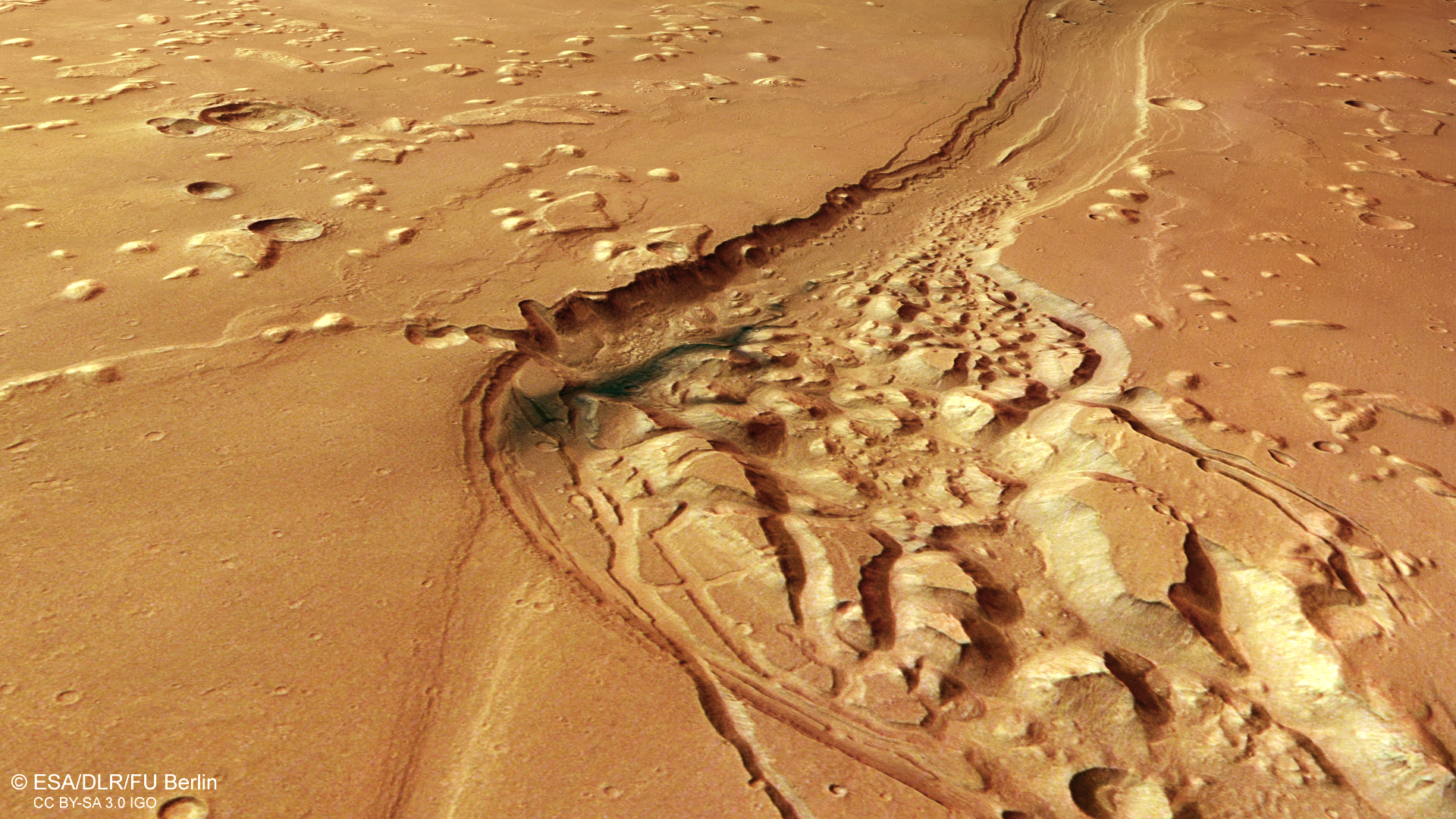 A dark orange patch of Mars, with a large, winding channel running from the bottom left to the right of the frame. The rest of the landscape is scattered with circular craters and uneven bumps, but is mostly smooth. A small scale bar at the bottom right indicates that north is to the left, and that a line approximately one tenth of the image width is 20 km long.