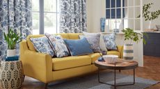 blue living room/open plan space with blue patterned curtains and cushions, yellow sofa, coffee table, footstool, rug