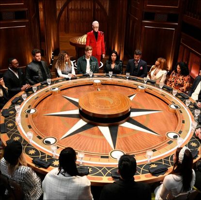 a group of people sit around a round table decorated with a star motif, as alan cumming stands among them wearing a red suit, in 'the traitors'