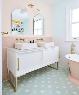 Pastel themed bathroom, blue and white geometric tiles, mixture of different tile shapes and color on walls, walls painted a range of pastel colors, large white vanity uniy with twin sinks and arch mirrors, wall light over mirrors, pink bathtub