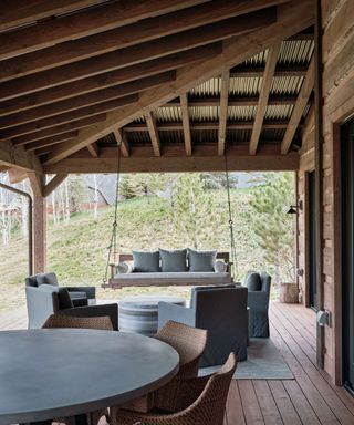 Outdoor terrace, porch area with dark wood paneled ceiling and metal roofing, seating area with armchairs and rounded ottoman, dining area with rounded table and chairs, hanging swing sofa seat from ceiling, dark wooden ceiling