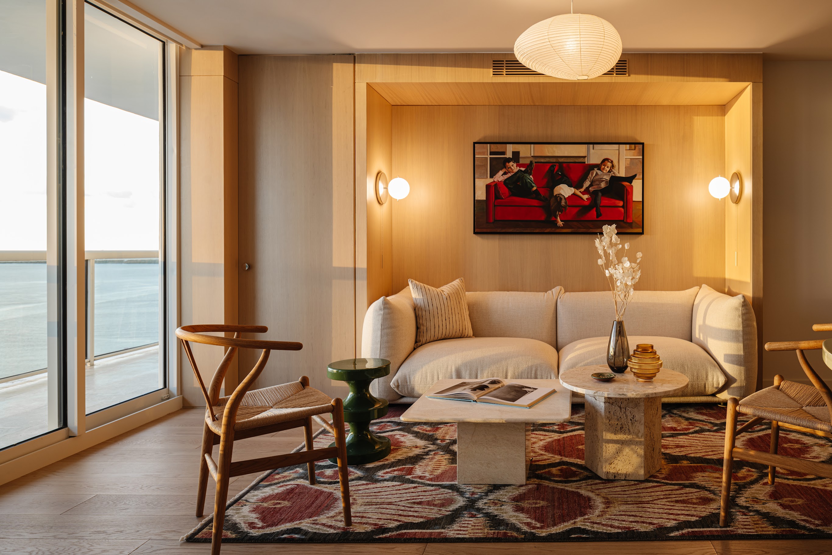 A naturally lit interior with a lounge sofa in a soft shape, wood-paneled walls, wood furniture along with marble accents, eccentric artwork, and a pendant paper lantern.