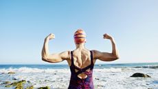 A woman wearing a bathing suit at the beach is flexing her arm muscles with her back to us and the beach in the background.