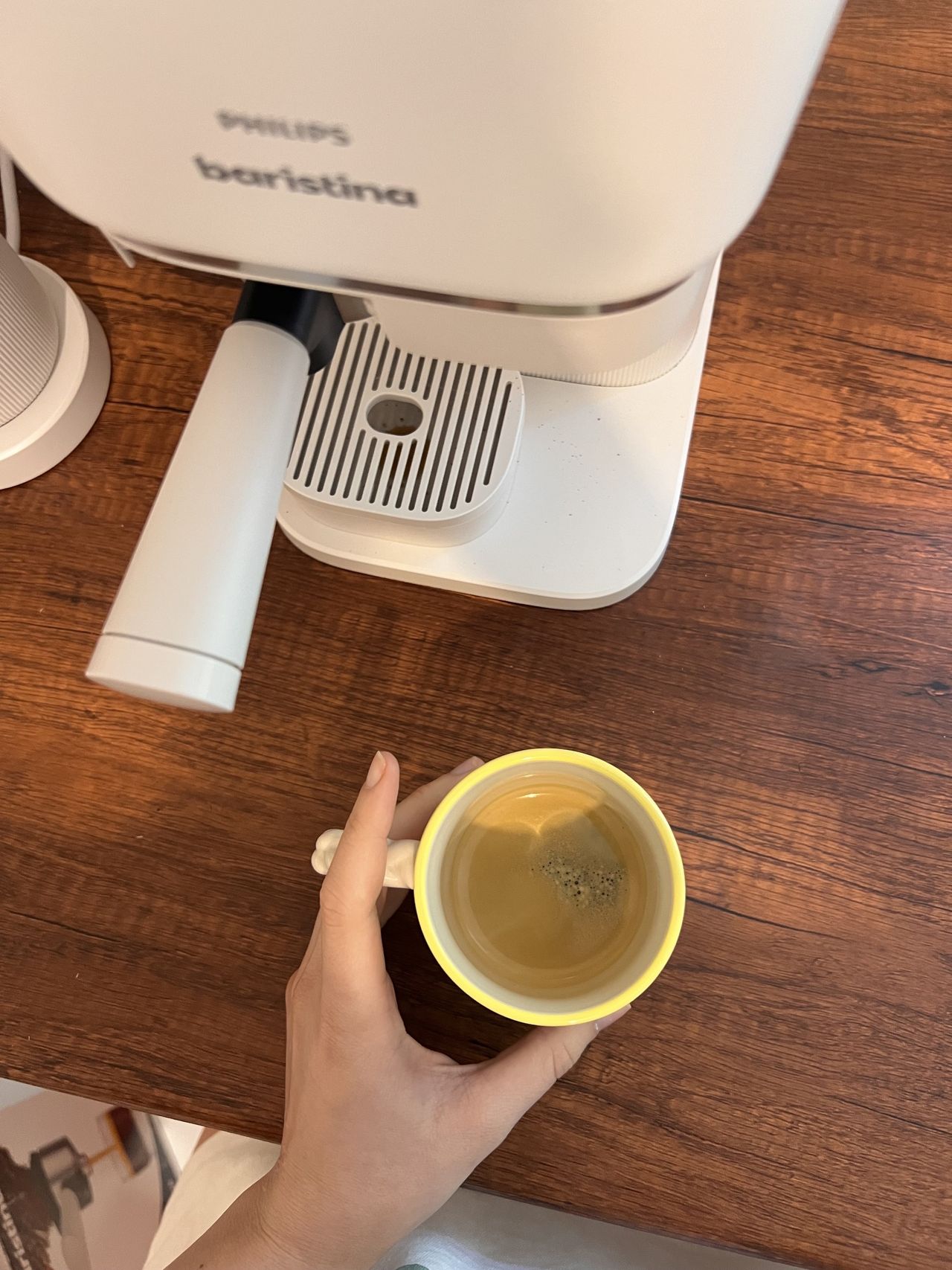 A hand holding a coffee mug on a wooden countertop that has an espresso shot in it. There is a white Philips Baristina also on the counter.