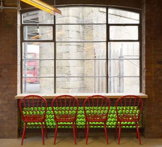 A table with red chairs in front of a large window with a round green pattern on the wall below it.