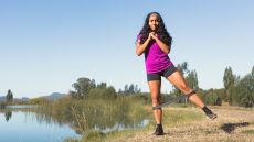 A woman performs banded hip abduction by a river on grass. She is standing on her right leg, her left leg straight and out to the side, and there is a resistance band around her calves.