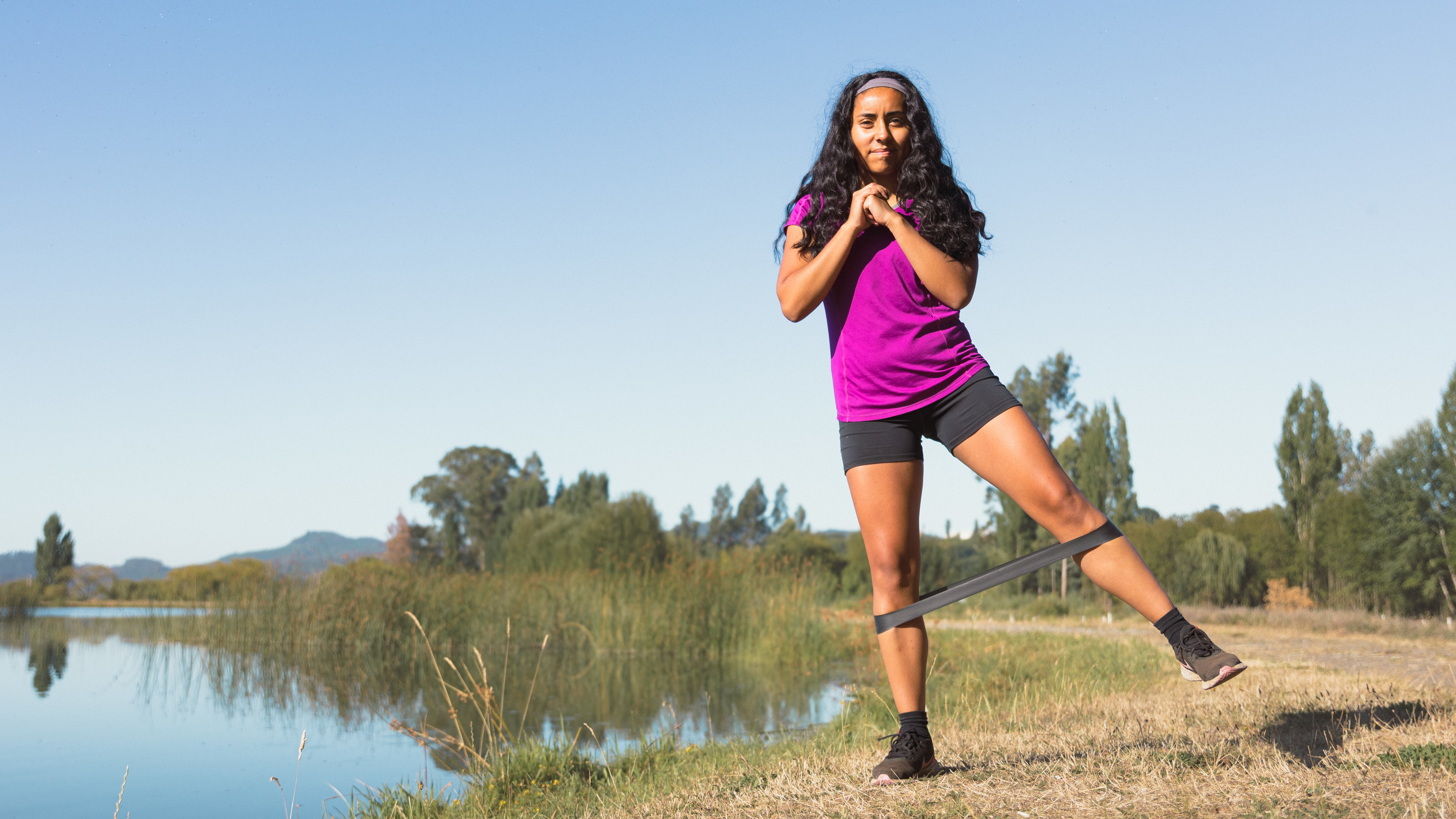 A woman performs banded hip abduction by a river on grass. She is standing on her right leg, her left leg straight and out to the side, and there is a resistance band around her calves.