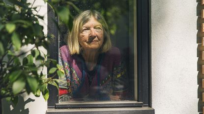 An unsure-looking older woman looks out a window.