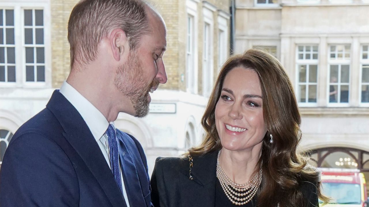 Kate Middleton has long, wavy brunette hair and wears a black jacket with a black top and a pearl necklace while gazing at husband Prince William&#039;s beard