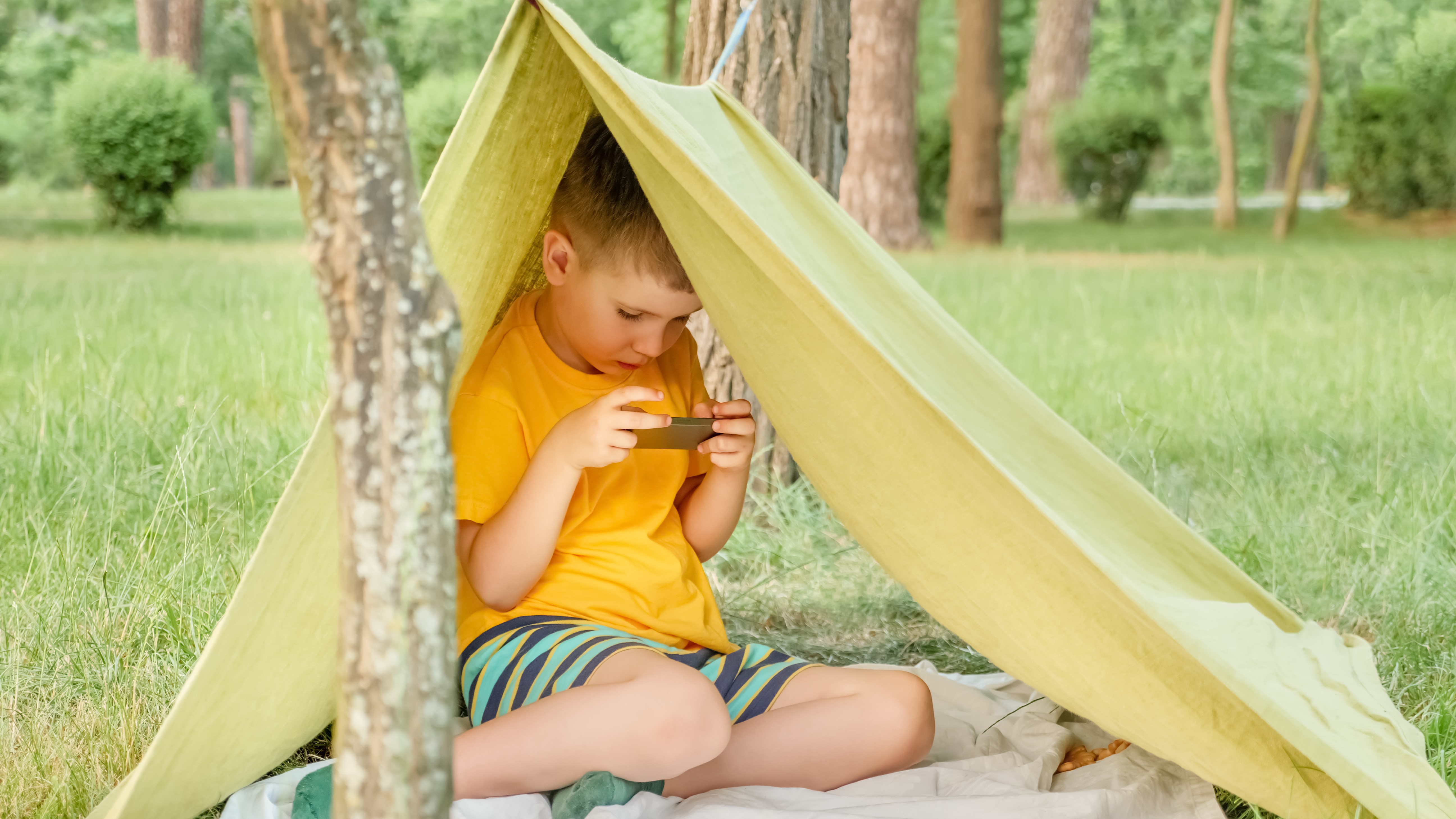 Un niño pequeño mira Netflix en su teléfono mientras está sentado en una carpa improvisada en el bosque