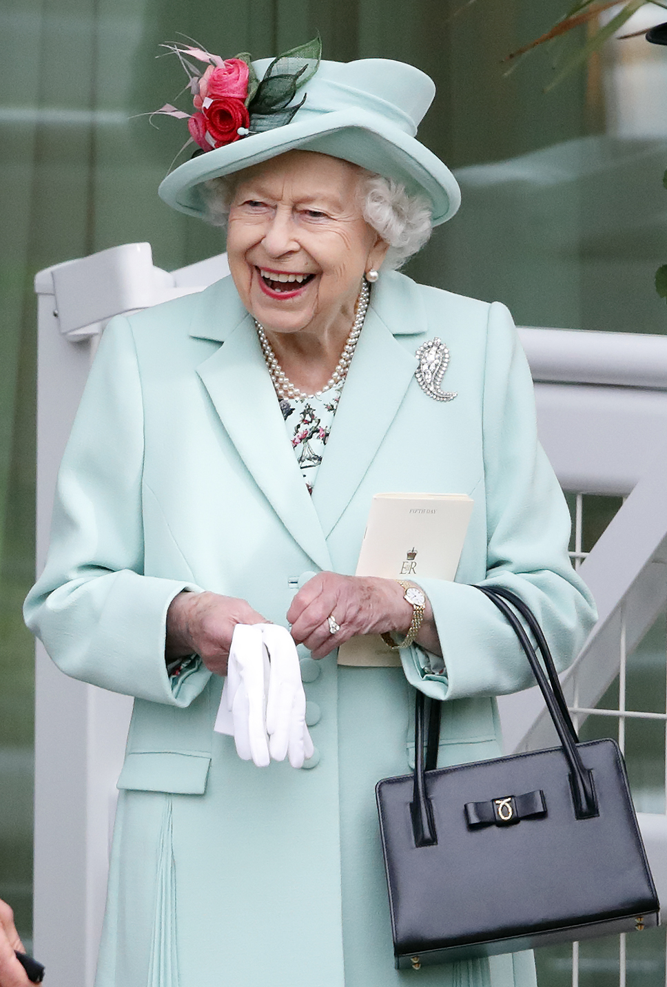 Queen Elizabeth standing in front of a white fence wearing a mint coat and matching hat, holding a black purse and white gloves and laughing