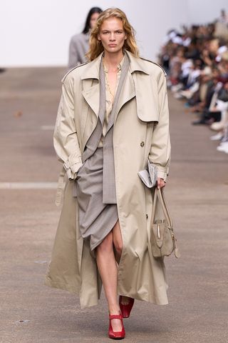 How to wear a suit is shown in a photo of a model on the runway of Stella McCartney's spring 2025 runway show wearing an oversized trench coat over a beige button-down shirt styled with a gray pinstripe blazer, a gray draped midi skirt, red Mary Jane heels, and a suede bowler bag