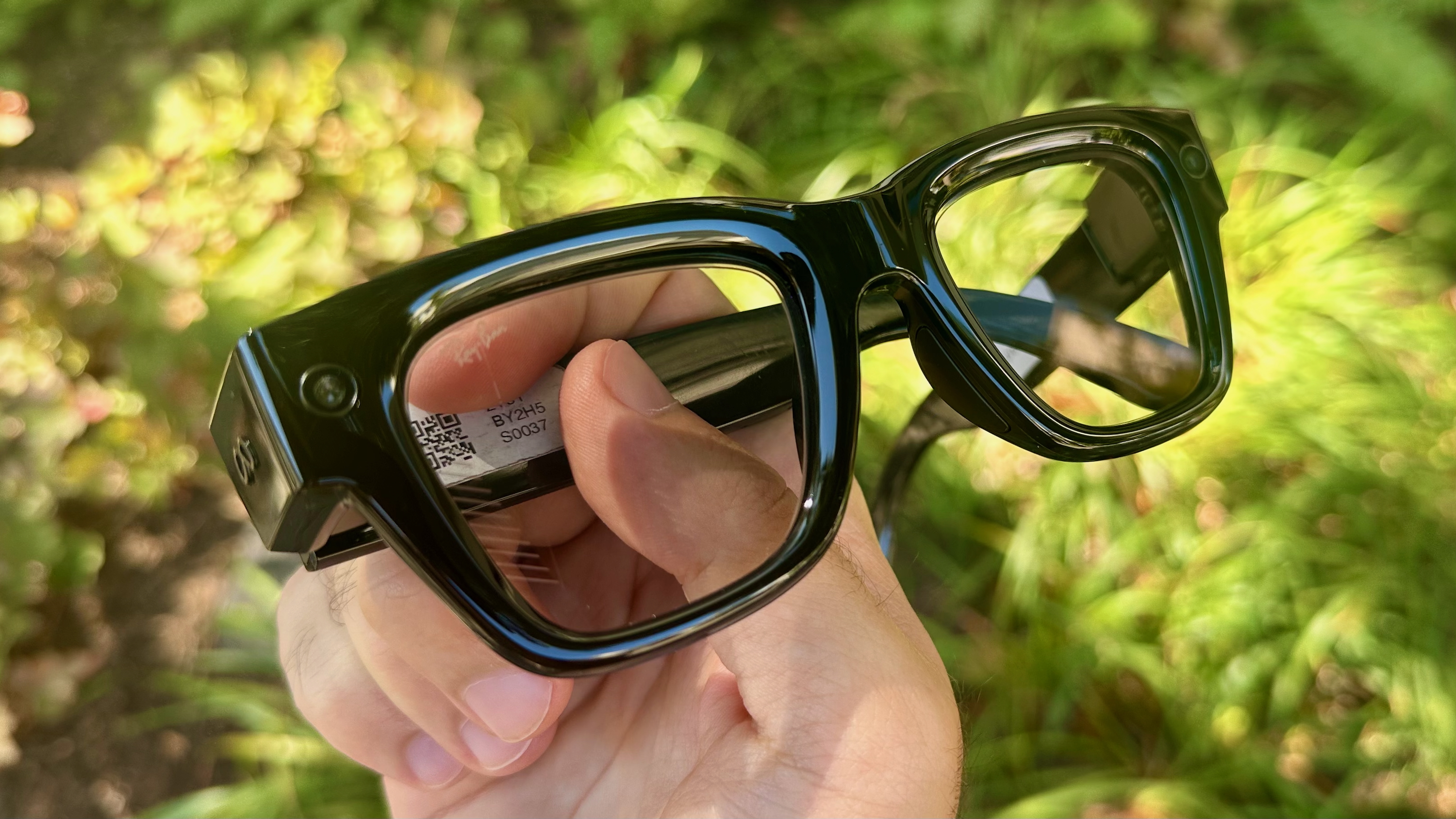 A close-up photo of the Meta Ray-Ban Display glasses held facing the camera to show the design, camera cutouts, and lenses.