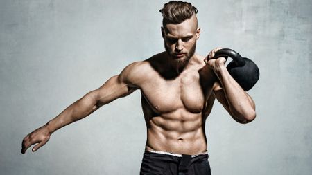 Man holding one of the best kettlebells up to his shoulder using left arm against a grey backdrop