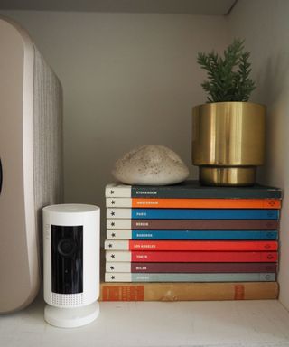 Ring Indoor Cam on a shelf beside a speaker and a stack of books with a plant