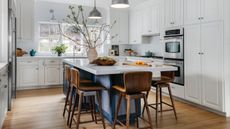 White kitchen with blue island, white countertop, hardwood floor, crhome pendants, wood and leather barstools, vase with foraged stems,