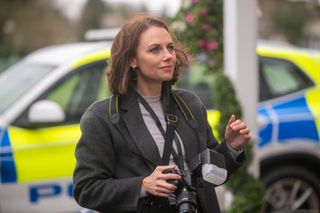 Jessica Ellerby as journalist Caitlin Dawson, wearing a long charcoal grey coat and carrying a professional-looking SLR camera around her neck