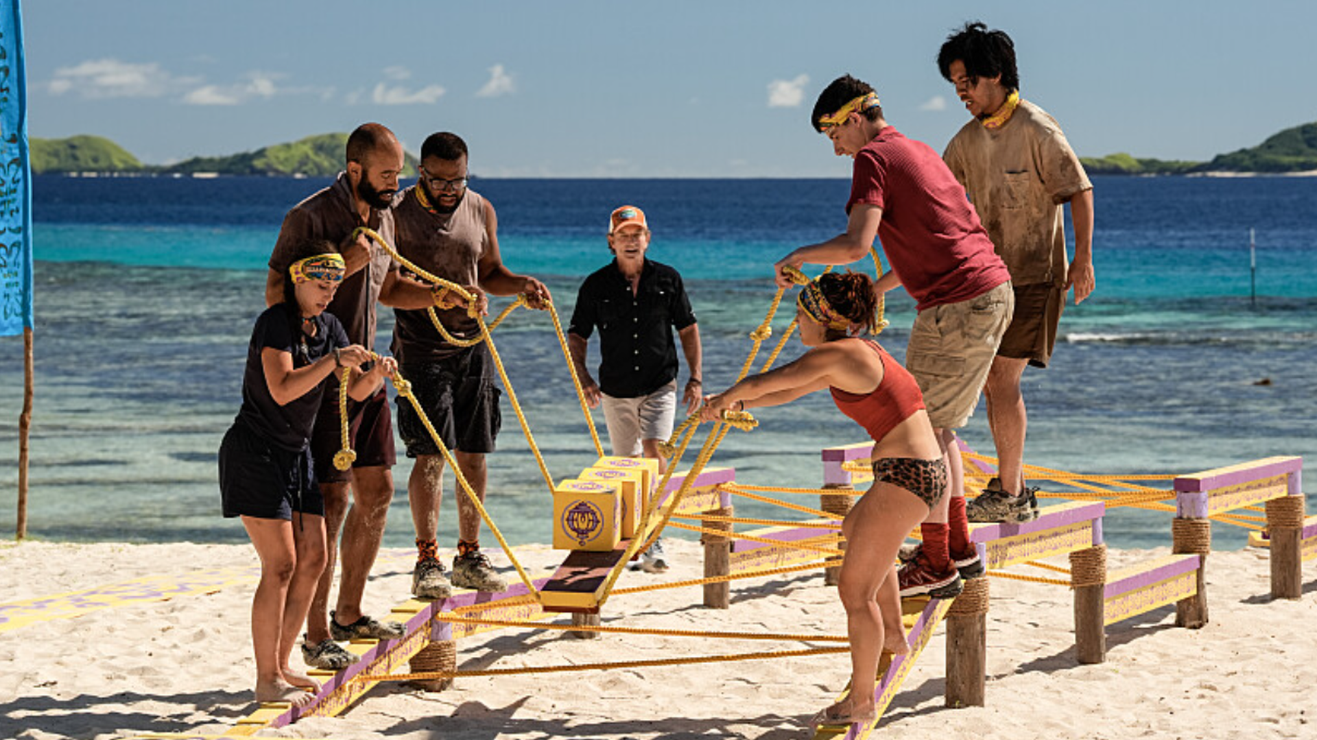 Sophi Balerdi, Nate Moore, Jawann Pitts, Savannah Louie, Rizo Velovic and Jason Treul competing in a challenge on the beach as Jeff Probst looks on in Survivor 49