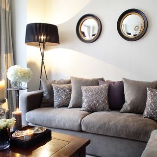 A white living room furnished with a grey sofa, wooden coffee table, tall floor lamp and round mirrors.
