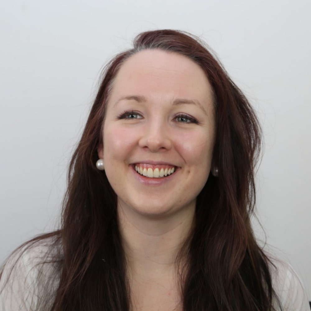 Amy Poulton profile picture. She is a white woman with long brown hair and is pictured smiling at the camera and against a gray background