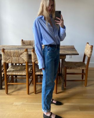 Fashion influencer Christie Tyler poses for a mirror selfie wearing a blue button-down shirt tucked into jeans with a black belt and black Mary Jane flats.