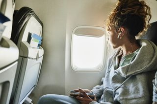 young woman sat on an aeroplane, looking out of the window and wearing earphones.