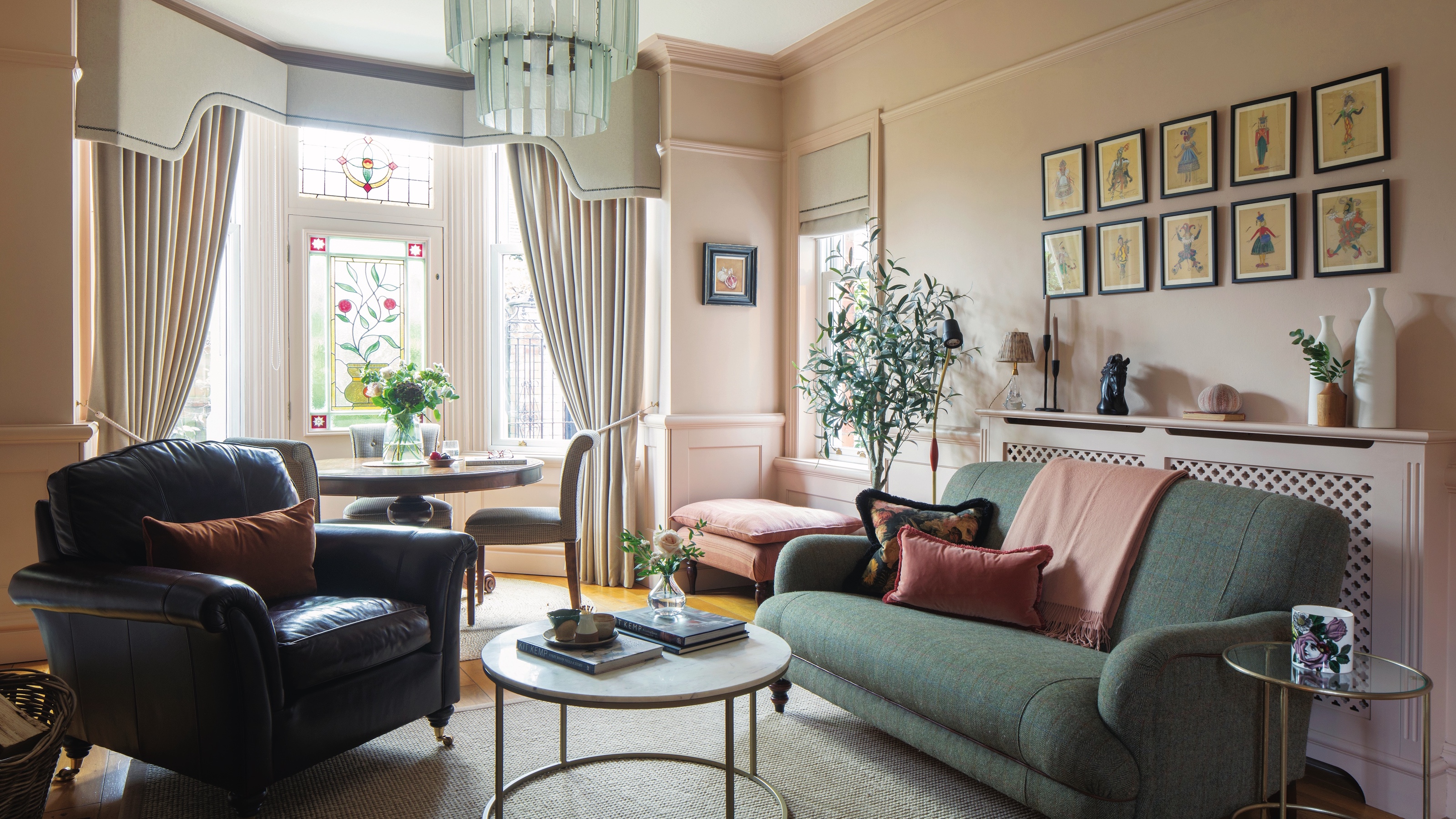 sitting room with blush pink walls, green sofa and armchair, with round coffee table and gallery of prints on wall.
